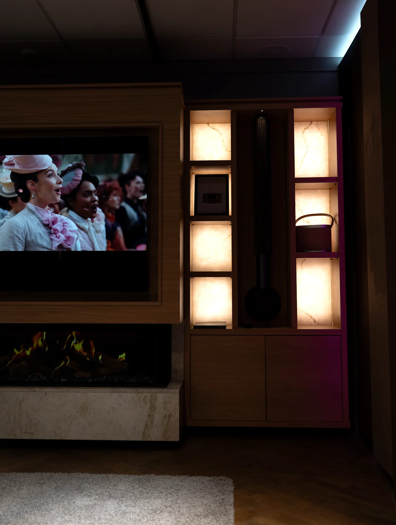 Backlit onyx alcoves and a wall-mounted loudspeaker in a darkened room, with a television and fireplace lit on the left.