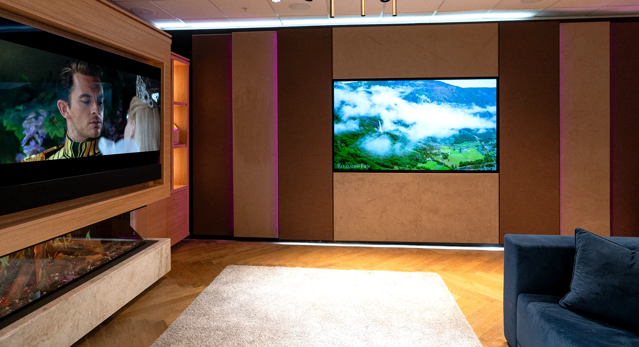 A large display set flush into a suede acoustic wall, with a second media wall and fireplace reflected along the left of the room.