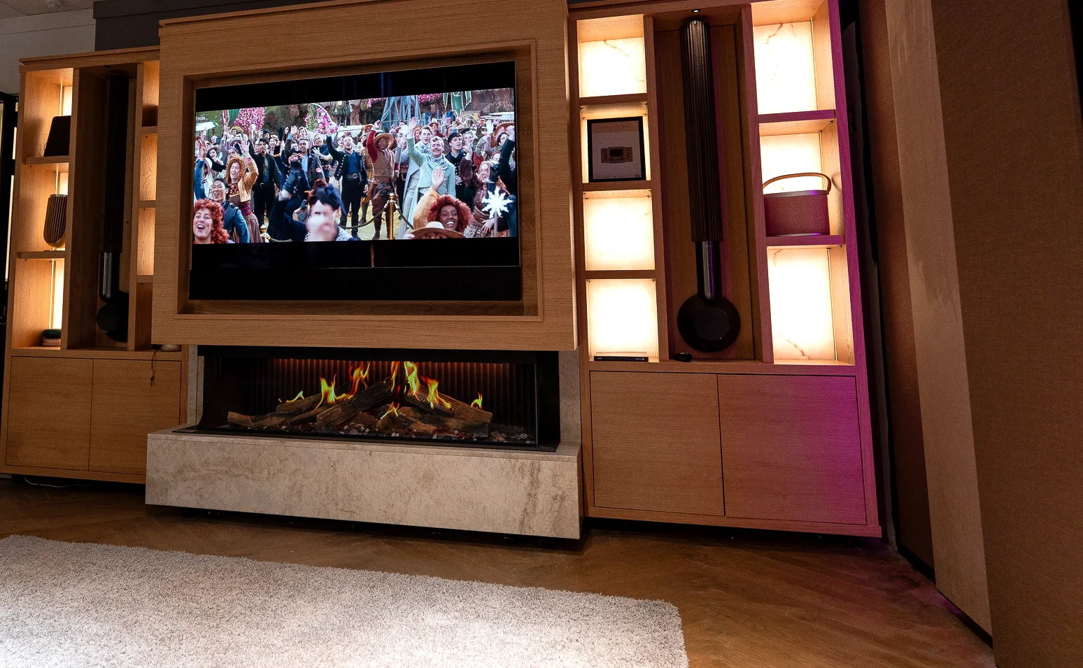 An oak media wall at an angle, television playing above a lit fireplace, with backlit onyx display niches on the right.