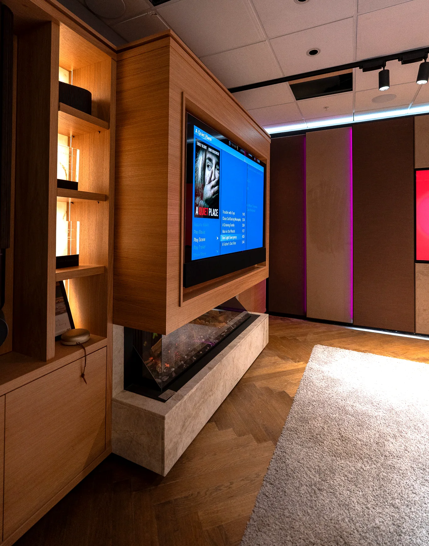 A media wall with an on-screen menu showing, above a linear fireplace, alongside lit oak shelving and a suede acoustic wall.
