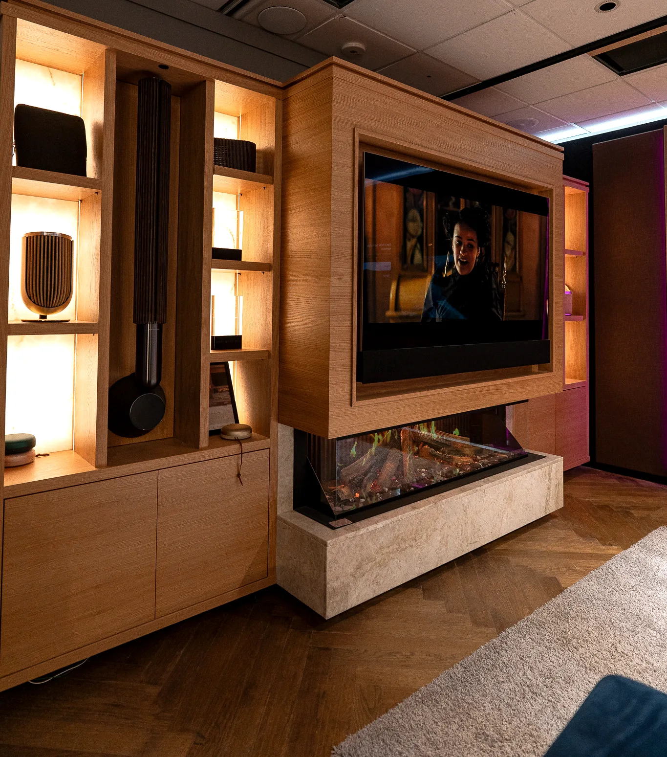 An oak media wall and fireplace beside a lit display cabinet holding loudspeakers, with a herringbone parquet floor.