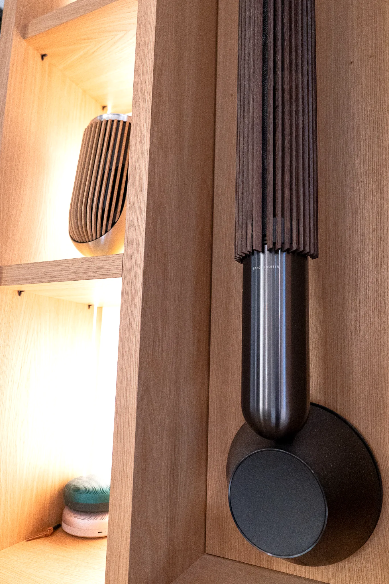 A wall-mounted Bang and Olufsen loudspeaker in oak cabinetry, with smaller speakers on the lit shelves beside it.